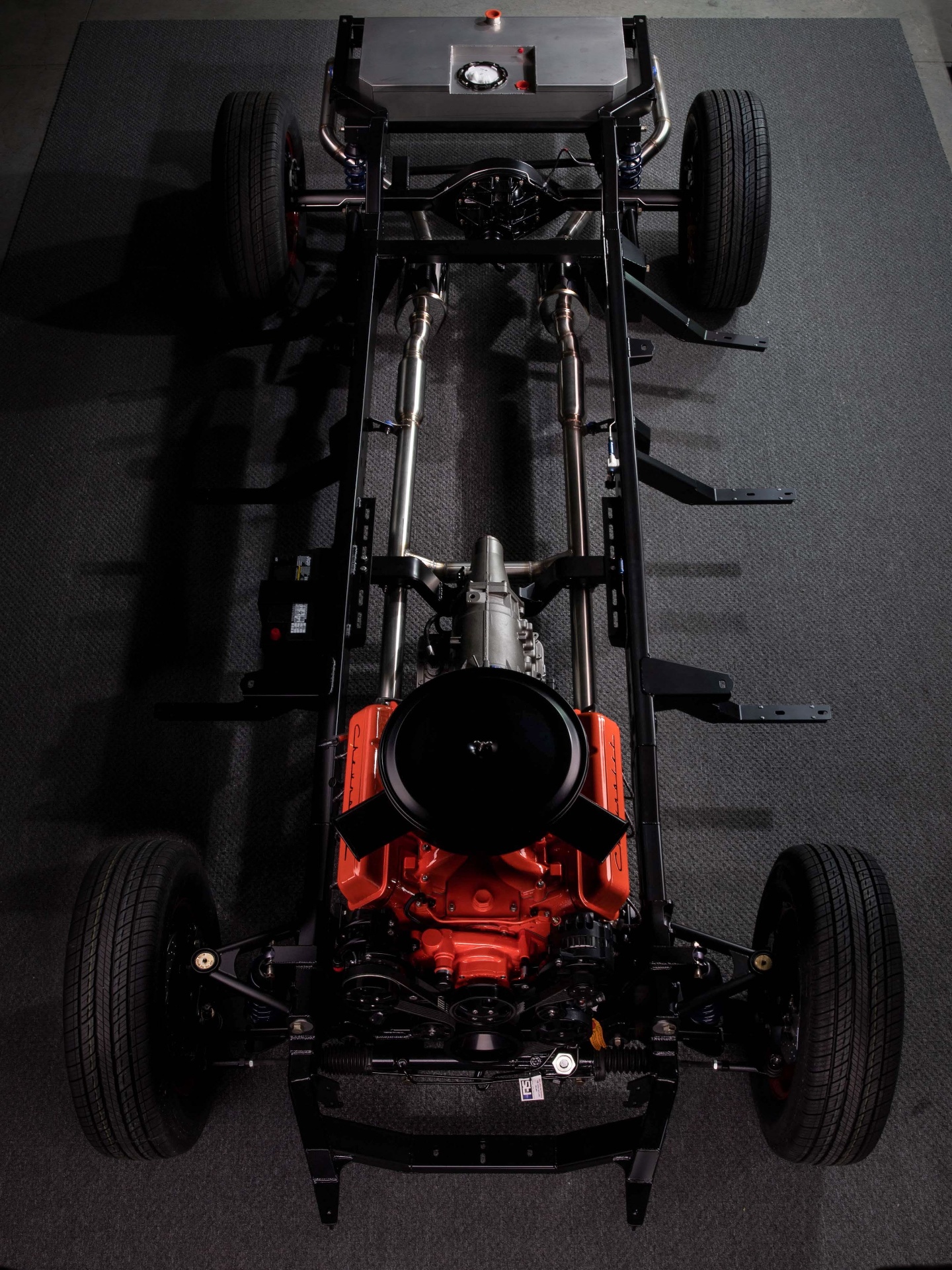 Rolling chassis, top-down — Roadster Shop SPEC frame in satin black powder coat, Don Hardy LS long-block already powder coated before assembly, Bowler 4L65E behind it, stainless exhaust routed along the rails.